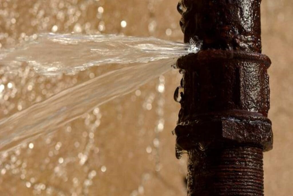 A rusty metal pipe with a significant leak, spraying water forcefully from its side against a blurred background.