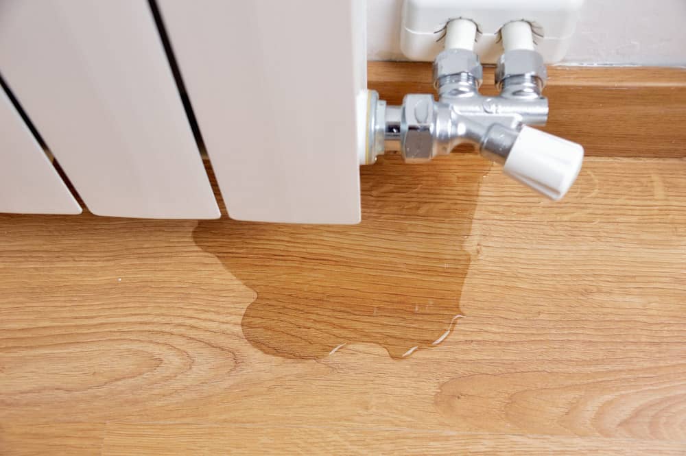 Water leaking from a white radiator onto a wooden floor, with the leak pooling near the radiator valve and pipes against a white wall.
