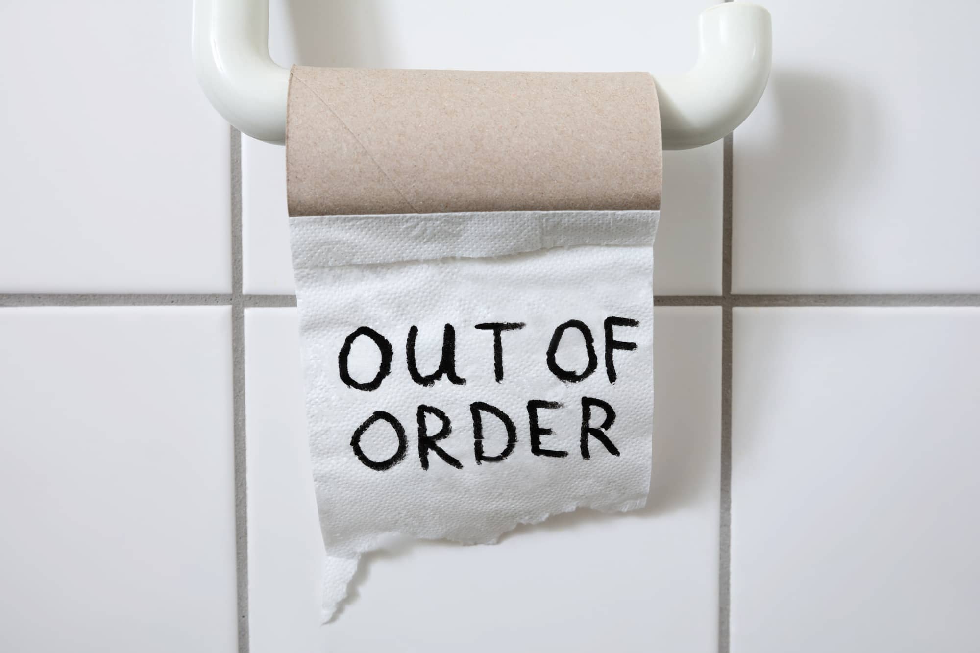 A toilet paper roll hung on a holder, with the last sheet hanging down and the words "OUT OF ORDER" written on it in black marker. The roll is empty, against a white tiled wall.